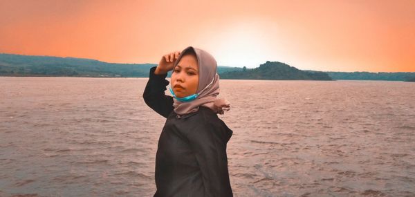 Young woman holding sunglasses while standing on beach against sky