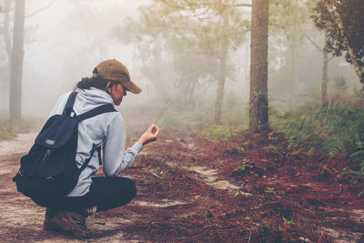Young woman picking twig while crouching in forest during foggy weather