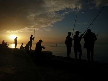 Silhouette people fishing at beach against sky during sunset