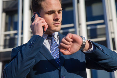 Low angle view of businessman talking on phone while checking time in city