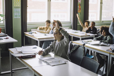 Boy with hand raised sitting at desk while attending lecture in classroom