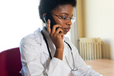 Competent young african american female medical practitioner answering phone call and using laptop while consulting patients remotely from office
