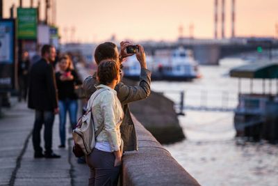 Man photographing while standing on street by lake during sunset