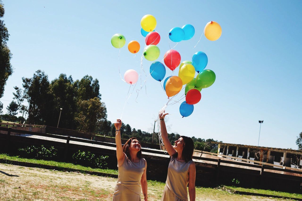 real people, balloon, two people, leisure activity, lifestyles, mid-air, togetherness, day, casual clothing, celebration, men, women, multi colored, fun, outdoors, helium balloon, young women, tree, happiness, sky, clear sky, young adult, bubble wand, adult, people