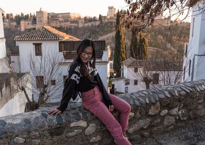 Full length of happy young woman sitting on retaining wall against buildings