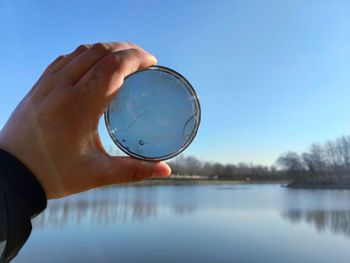 Cropped hand of person holding mirror with reflection over lake against sky