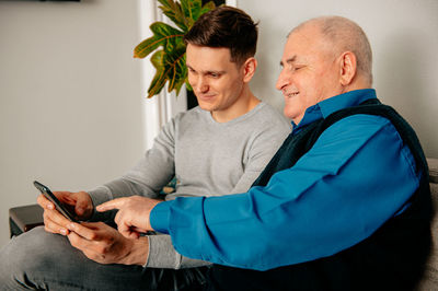 Happy young man with grandfather using smart phone at home