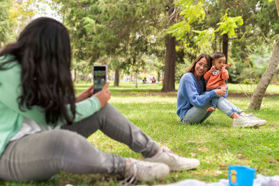 Young female taking photo on cellphone of cheerful mom raising baby while sitting on lawn in park