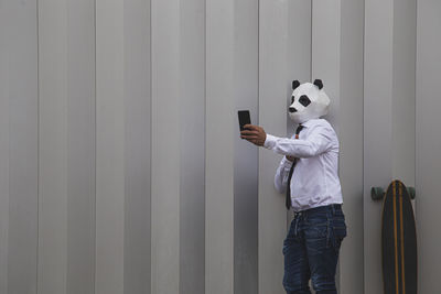 Man in white shirt, tie and panda mask taking smartphone selfie against wall