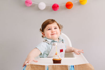 Cute baby boy with birthday cake sitting on high chair at home