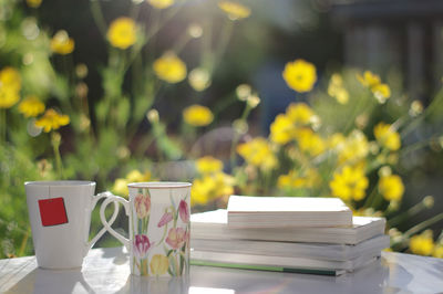 Close-up of books with mugs on table against flowers