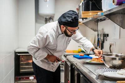 Concentrated male cook in uniform standing at metal counter in kitchen of cafe and preparing delicious dish with salmon fish