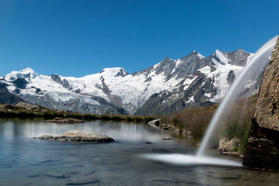 Scenic view of waterfall by lake against snowcapped mountains