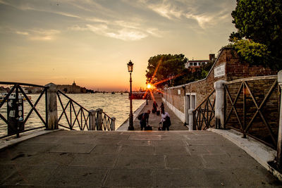 People on street by illuminated city against sky during sunset