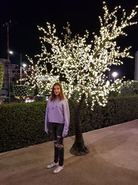 Portrait of young woman standing by illuminated tree at night