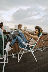 Full length of cheerful woman holding hand of male friend sitting on chair at rooftop