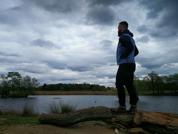 Full length of man standing on dead tree by river against cloudy sky