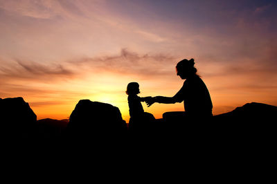 Silhouette mother and daughter holding hands against orange sky