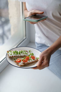 Woman takes a photo on cellphone hold plate with rye crisp bread with creamy cheese and micro greens