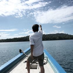 Rear view of young man with hand raised standing on boat in lake against sky