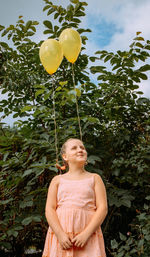 Girl in a pink dress stands on the street. yellow balloons are tied to her braided hair.