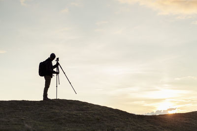 Side view of silhouette hiker fixing tripod on hill against sky during sunset