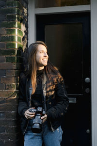 Concentrated young ethnic female traveler with long dark hair in casual outfit leaning on brick wall of aged building and watching photos on digital camera on sunny day in amsterdam