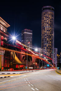 Light trails on road by illuminated buildings against sky at night