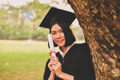 Portrait of young woman in graduation gown holding certificate while standing by tree at park