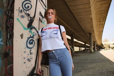 Portrait of young woman blowing bubble gum while standing under bridge