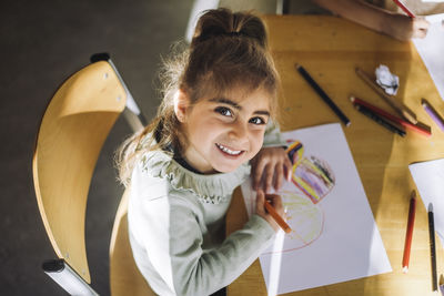 High angle view of smiling girl drawing with crayon on paper while sitting at bench in classroom
