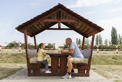 Mature man and girl eating food in wooden picnic table at parking lot