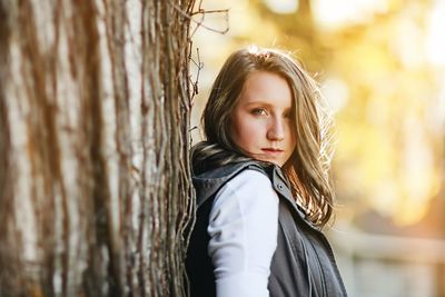 Portrait of beautiful young woman standing against tree trunk