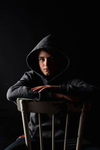 Portrait of adolescent boy in hoodie sitting on a chair in dark room.