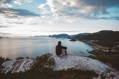 Rear view of man sitting on rock by sea against sky