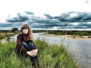 Portrait of woman sitting on grassy field by river against sky