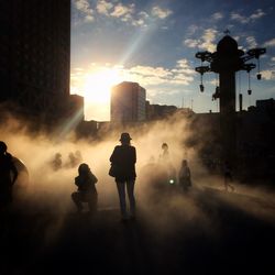 Silhouette people amidst smoke on footpath in city against sky during sunset