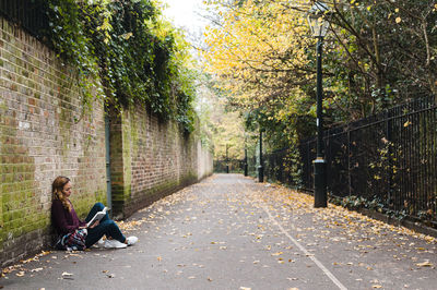 Woman reading book while sitting by brick wall on footpath during autumn