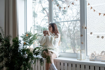 Woman in warm white winter sweater sitting on the window at home at christmas eve holding cup