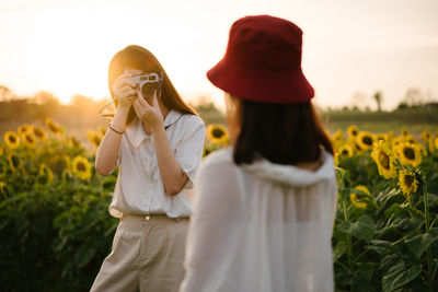 Young woman photographing female friend standing at sunflower farm during sunset