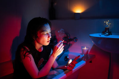 Young woman with cigarette smoking, scrolling social media on smartphone while sitting in bathtub