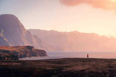 Remote side view of female explorer walking along rocky coast near sea on background of sunset sky and highlands on gran canaria