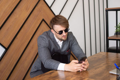 A young man in a suit and sunglasses is sitting at a desk in the office and looking at the phone
