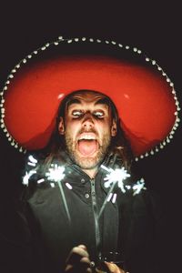 Low angle view of young man wearing hat holding sparklers while standing against black background