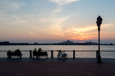 People sitting on benches by river against sky during sunset