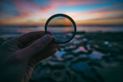 Cropped image of hand holding lens at beach during sunset