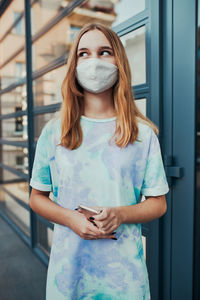 Portrait of young woman girl standing at store front downtown holding smartphone. girl wearing mask