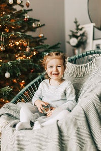 A little girl is sitting in an armchair and smiling against the background of a christmas tree 