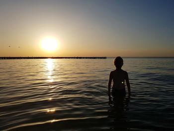 Boy standing in sea against sky during sunset