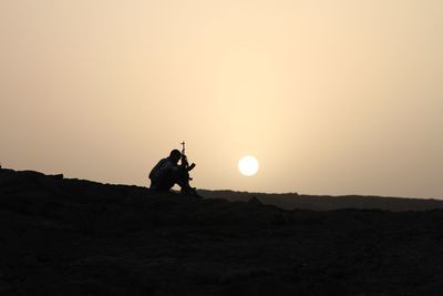 A security guard with a gun sits on the ground at sunrise.